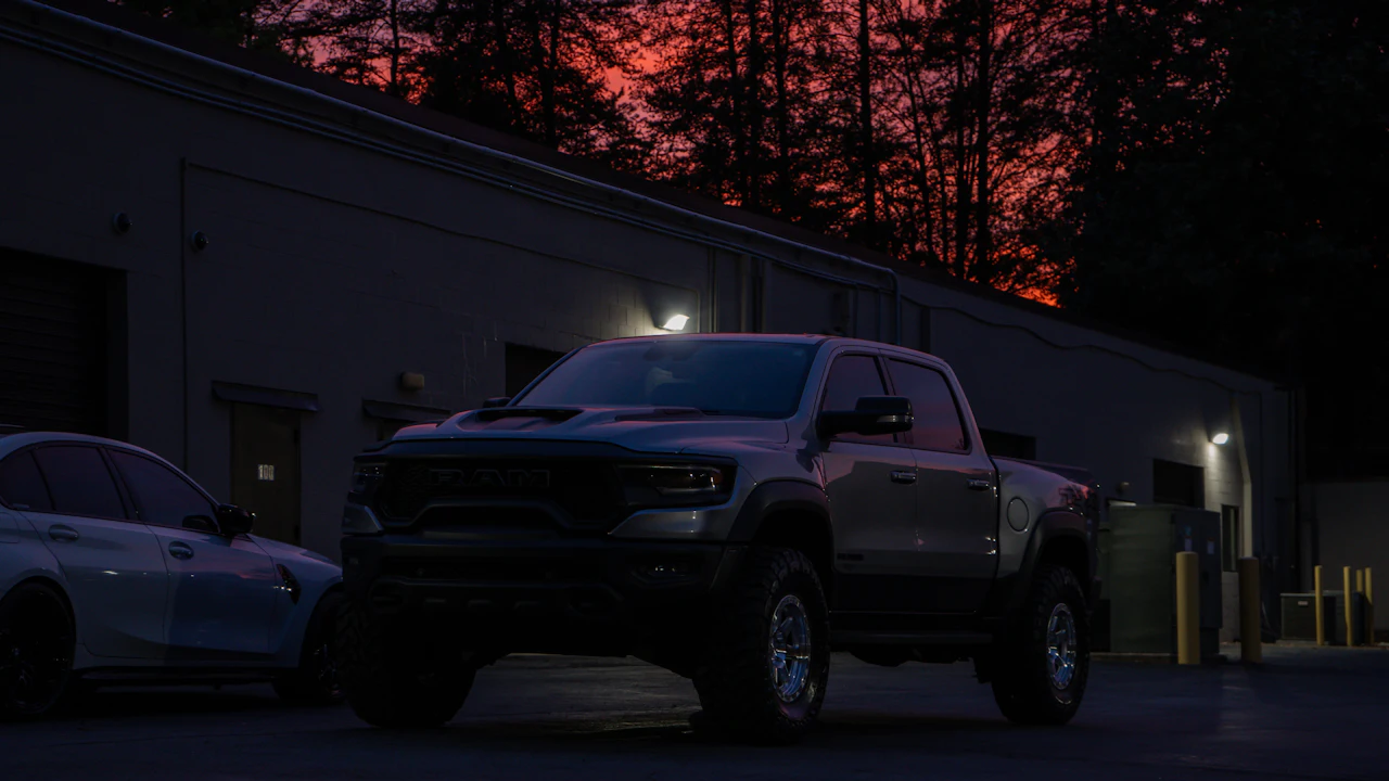 A large blacked-out American pickup truck parked outside an industrial unit at dusk, a deep red sunset behind bare trees