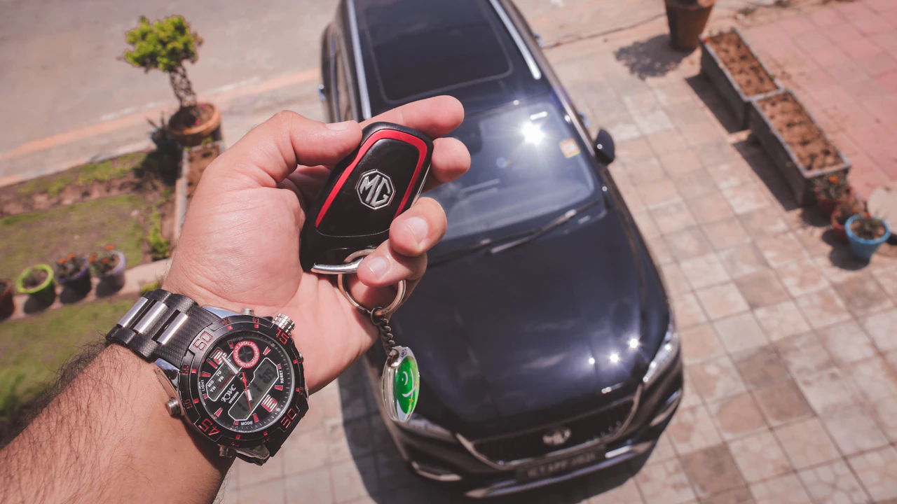 A hand holding a car key fob in front of a parked car on a driveway