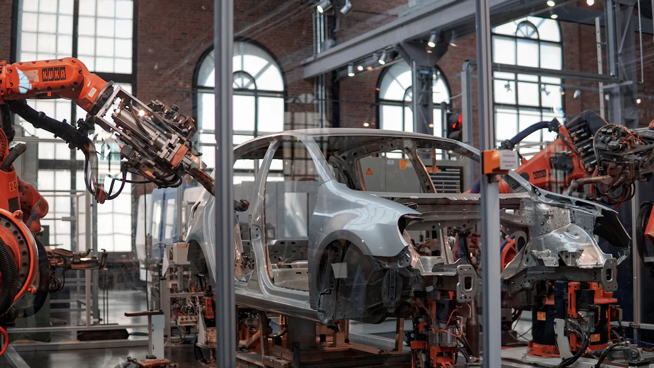 A bare silver car body-in-white standing on a production line in a brick-walled factory hall, orange industrial robot arms reaching toward it from both sides and a second shell visible further down the line.
