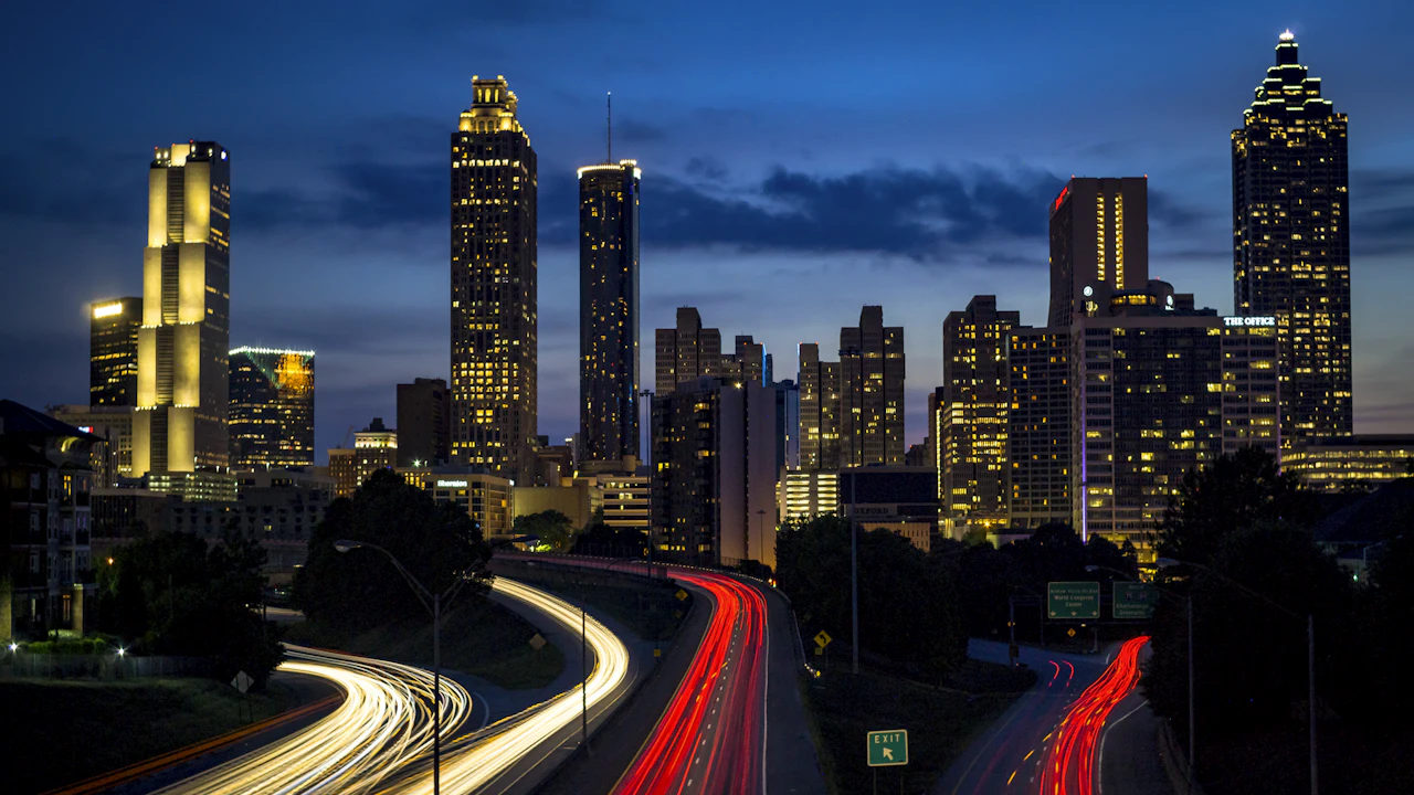 The Atlanta skyline at dusk above a freeway interchange, headlight and tail-light trails curving through the frame