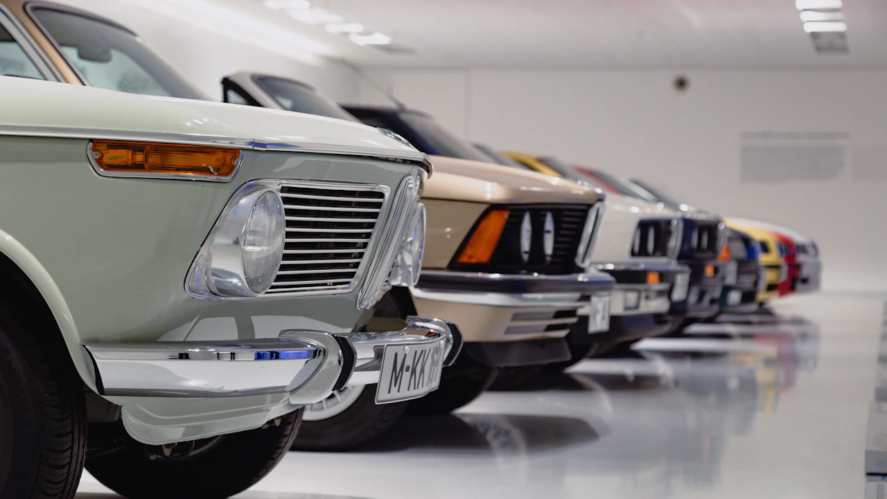 A line of a dozen older BMW saloons and coupes parked nose-out on a polished white floor under strip lighting, receding into the distance. A pale green car with round headlights and a chrome bumper fills the foreground, a cream coupe stands behind it, and the cars beyond fade into red, yellow and blue in the depth of the room.