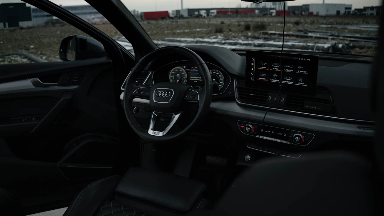 An Audi interior photographed from the driver&rsquo;s door, digital instrument cluster and central screen lit, an industrial yard beyond the glass