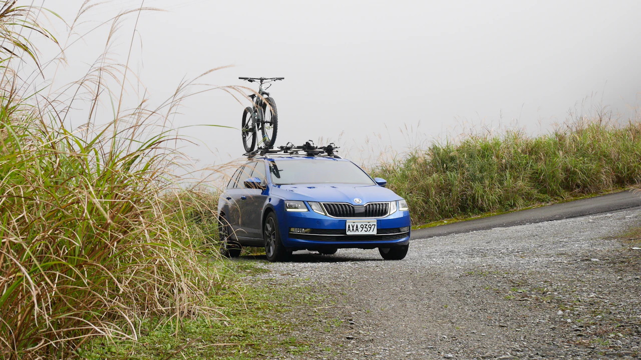 A blue estate car with a mountain bike on the roof rack, parked on a gravel mountain road with tall grass and low cloud behind it