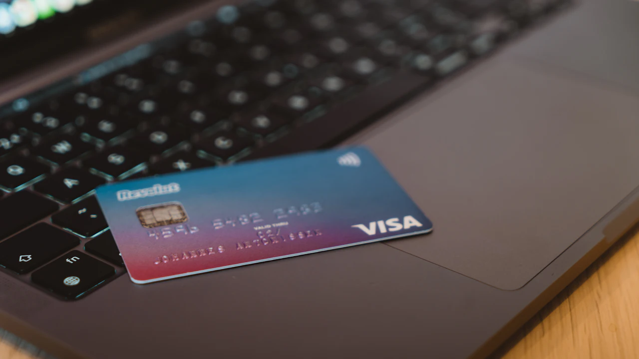 A blue-and-red gradient Visa debit card resting at an angle on the dark keyboard of an open laptop, the chip and embossed number catching the light.