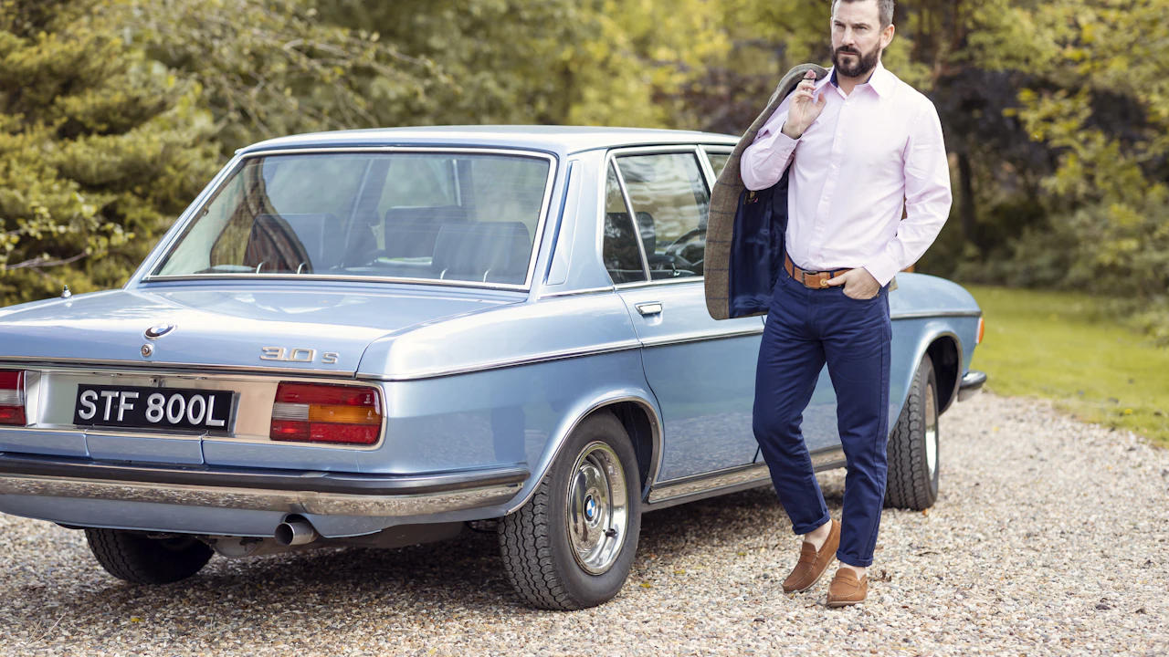 A man with his jacket over one shoulder walking away from a pale blue classic saloon parked on a gravel drive