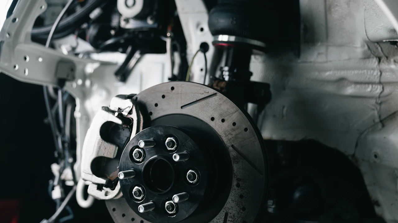 The front corner of a car with its wheel removed in a workshop: a drilled and slotted brake disc on a black hub carrying wheel studs, a silver brake caliper clamped over the disc on the left, and above the disc a suspension strut topped by an air spring, set against white-painted inner wing panels and wiring.