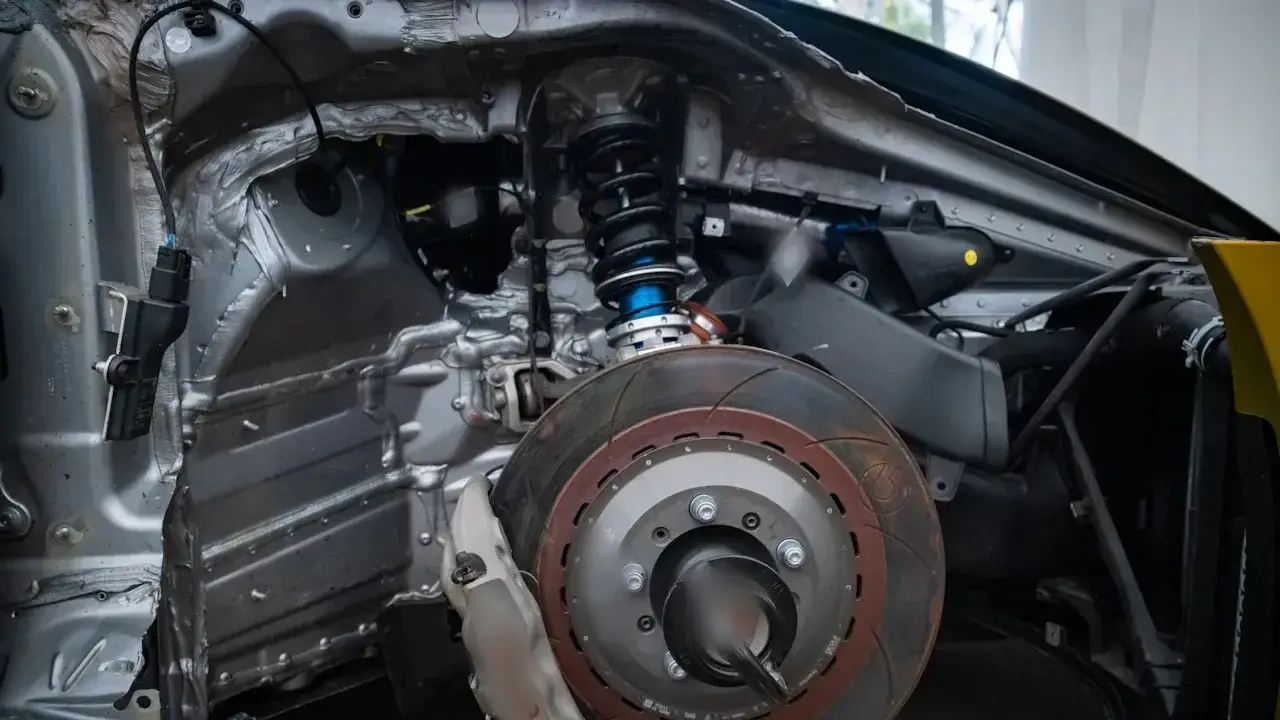 A car's front corner with the wheel removed: a slotted brake rotor, the brake caliper, wheel studs and the coil-over strut above.