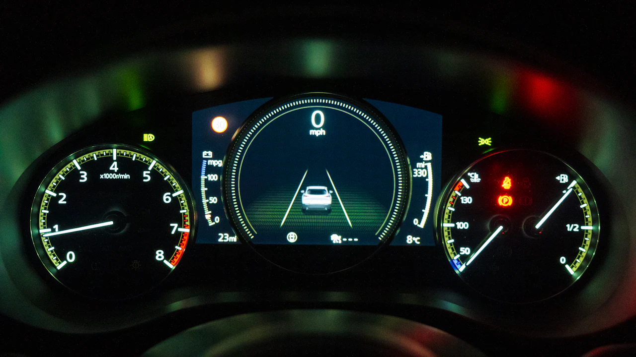 A car’s illuminated instrument cluster at night: a rev counter on the left, a central digital display reading zero miles per hour with a lane graphic, and a speedometer with fuel gauge and warning lights on the right.