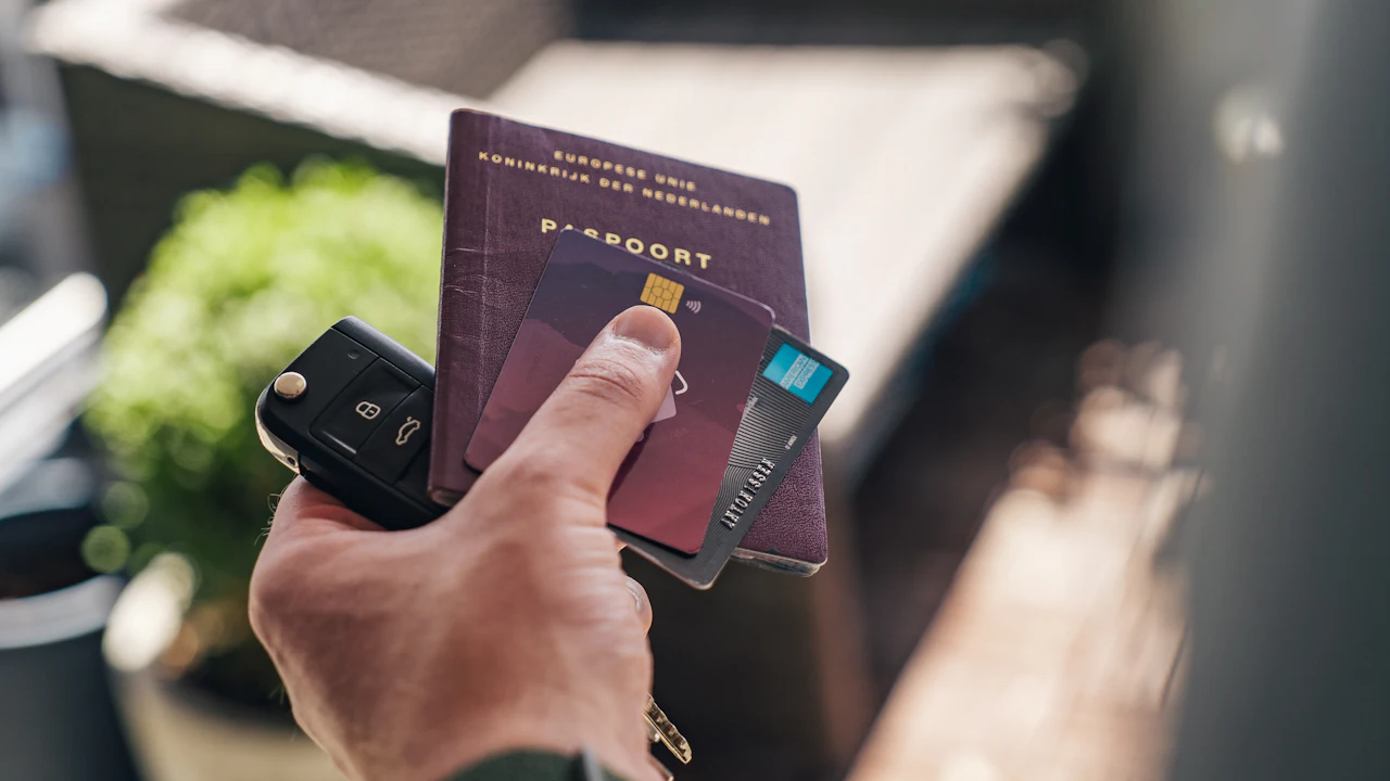 A hand holding a black car key fob together with a burgundy passport and two payment cards, a blurred garden behind.