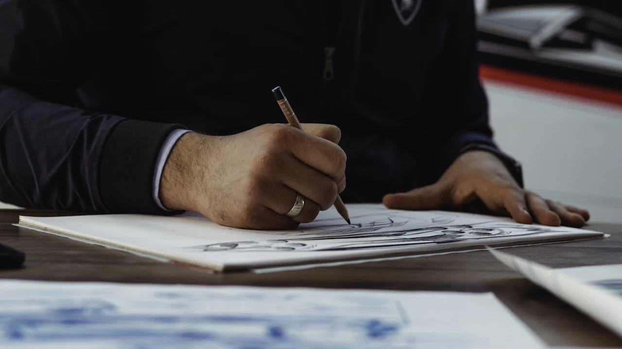A person in a dark suit writing with a pen on printed paperwork at a wooden desk, one hand resting flat on the sheet, more documents to the side.