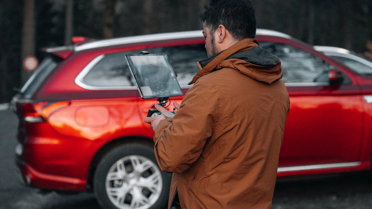 A man in a brown jacket holding a tablet, photographing and inspecting a red SUV parked on a gravel lot with dark trees behind.