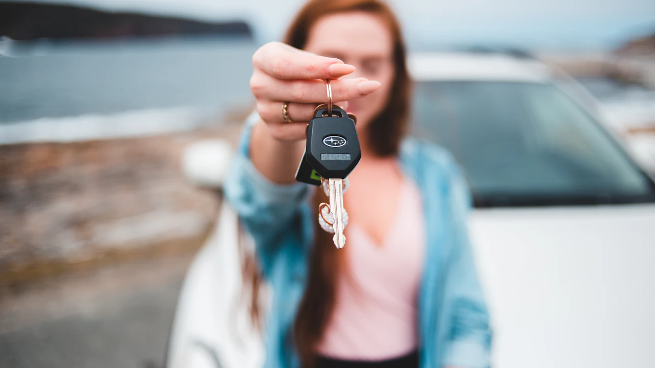 A young woman holding out a car key toward the camera beside a white car, a rocky coastline behind her