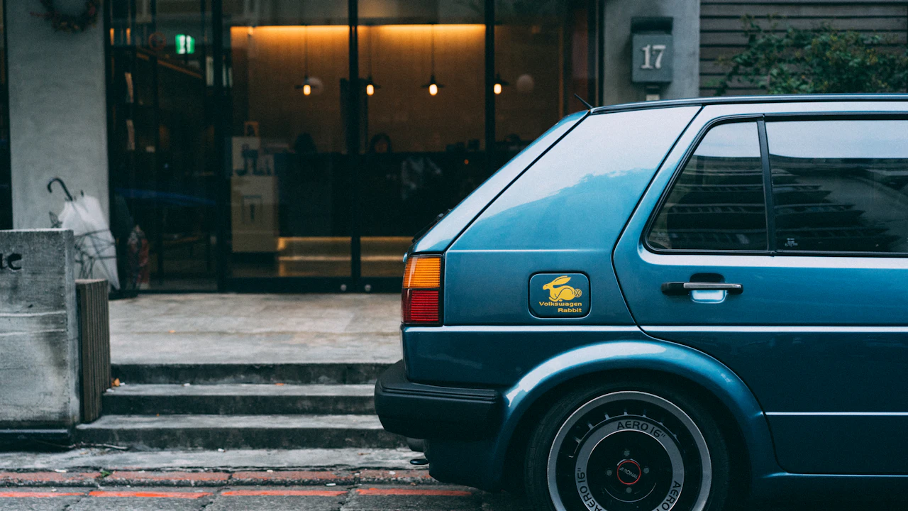 A blue Volkswagen hatchback parked at the kerb outside a lit shopfront numbered 17, seen from the rear three-quarter, its upright rear glass, tail light and alloy wheel in view.