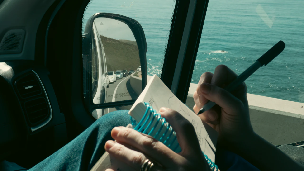 A person in a car holding a small spiral notebook and a pen, the wing mirror beside them showing a coastal road and traffic behind.