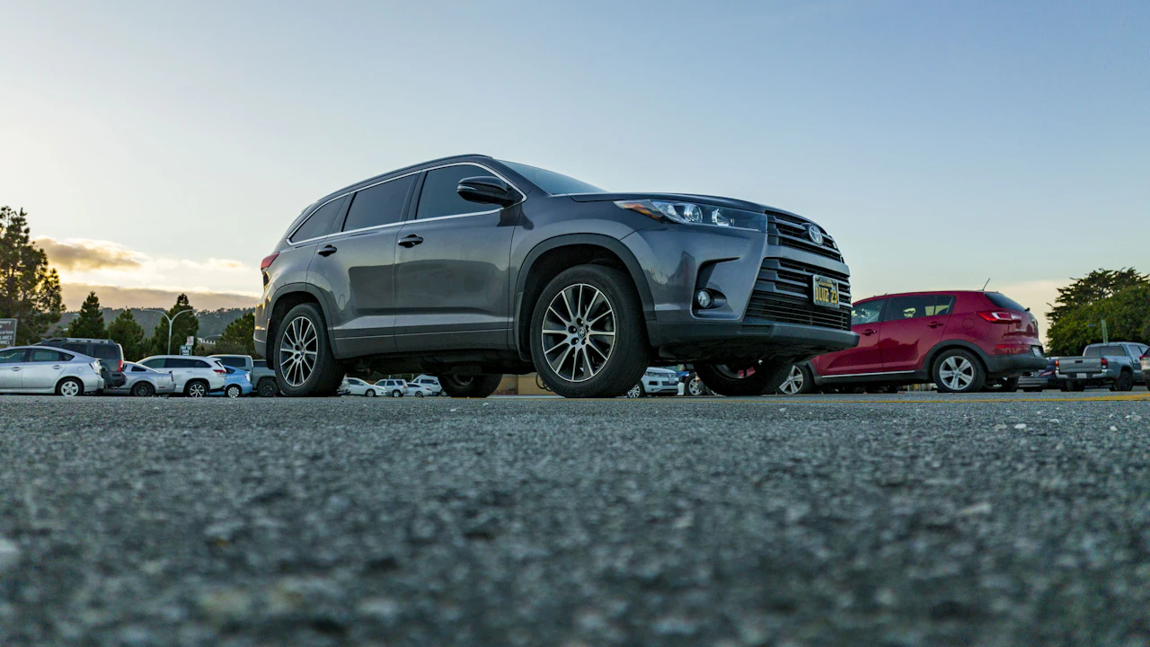 A grey Toyota Highlander SUV parked front three-quarter on a lot at dusk, other cars and a red hatchback around it, hills on the horizon.