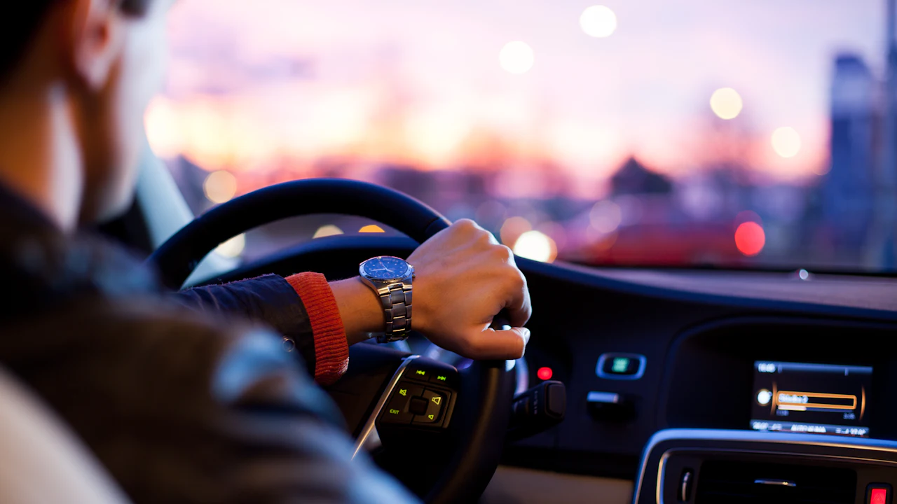 A driver's hands on a steering wheel seen from the passenger side, the road ahead through the windshield