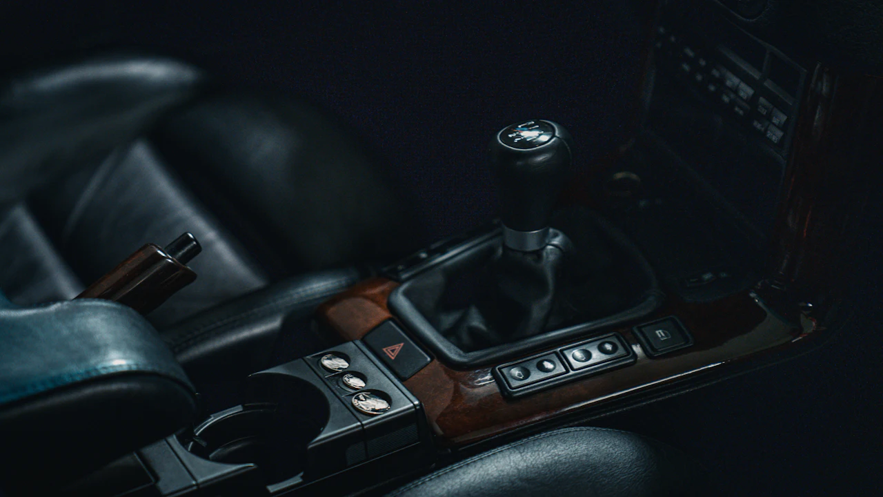 A manual gear shifter and centre console in a dark car interior with wood trim, a row of switches below the shifter and a handbrake to the left.