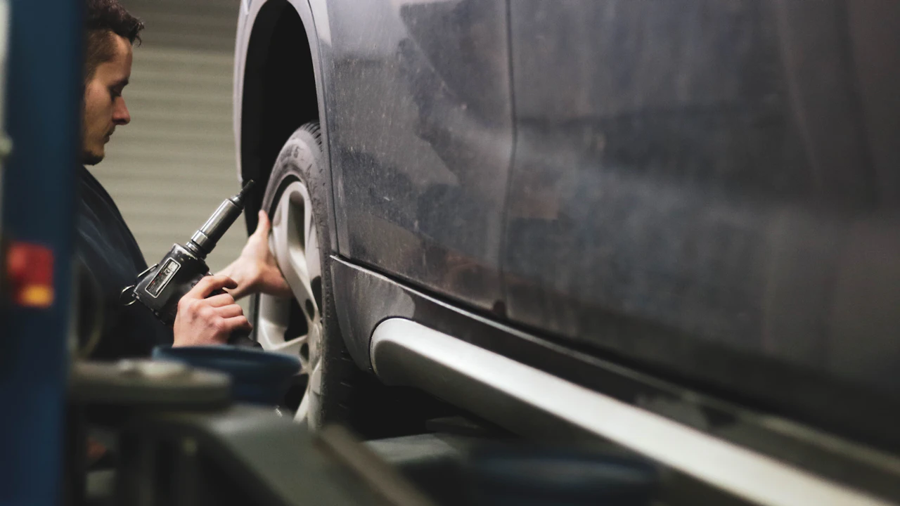 A technician in dark overalls crouches at the front wheel of a dusty dark-grey SUV raised on a two-post lift in a workshop, holding a cordless impact gun to the wheel nuts. The car’s side sill and running board run away to the right of the frame, and a blue lift arm crosses the bottom edge.
