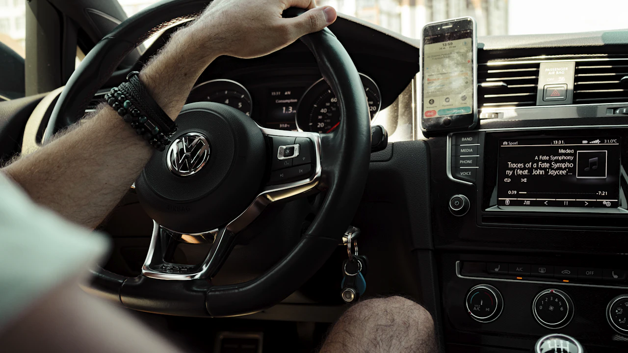 From the passenger side, a driver’s hand rests on a Volkswagen steering wheel while a phone clamped to the dash vent shows a map app, the centre screen and climate controls glowing below.