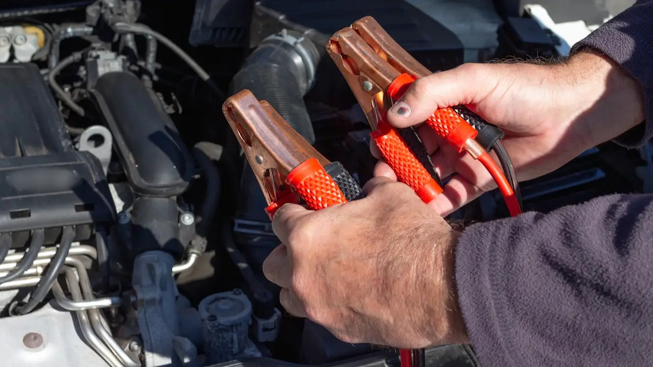 Hands holding the red clamps of a set of jumper cables over an open engine bay; engine-cover lettering blurred.