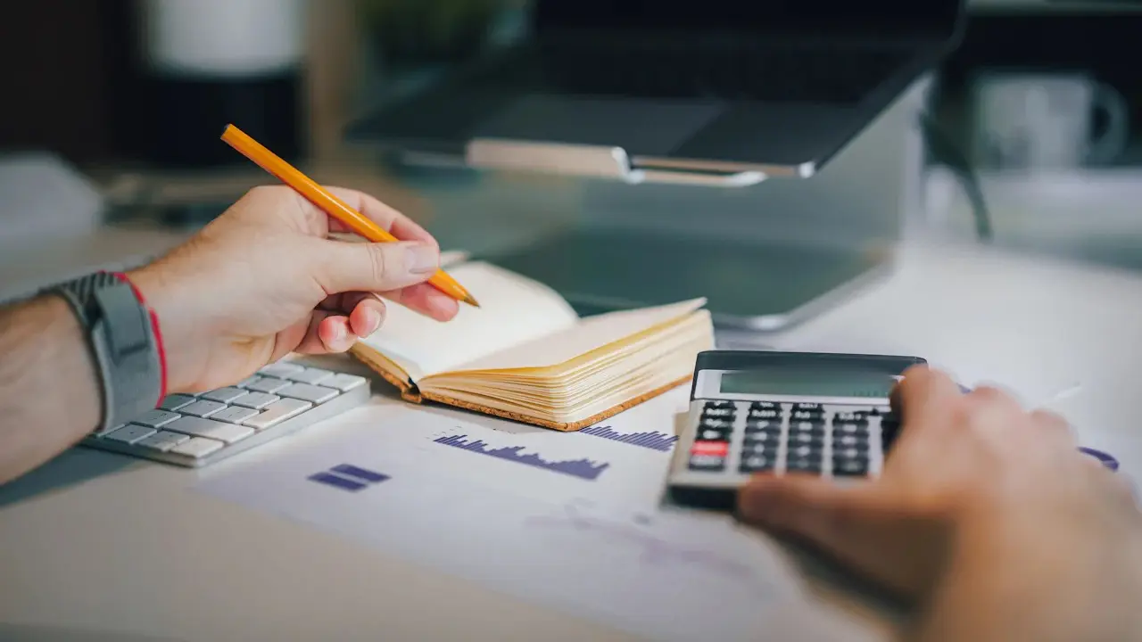 Hands writing in a notebook beside a calculator and keyboard at a desk.