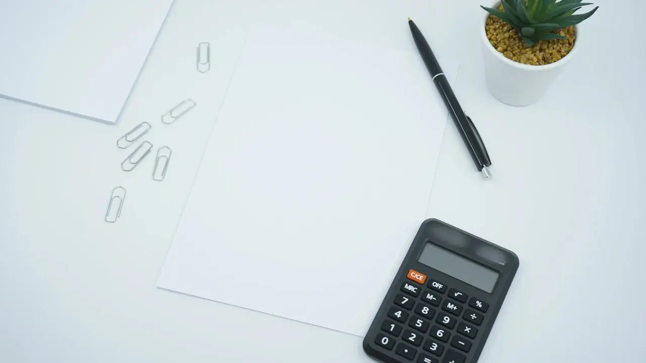 A calculator, a pen, paper clips and blank sheets on a white desk; the calculator's brand blurred.
