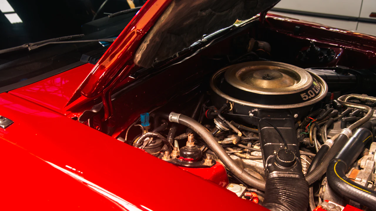 A red car with its bonnet propped open in a workshop, showing a V8 engine under a round chrome-lidded air cleaner, a ribbed intake duct, braided and rubber hoses, and wiring along the inner wings.