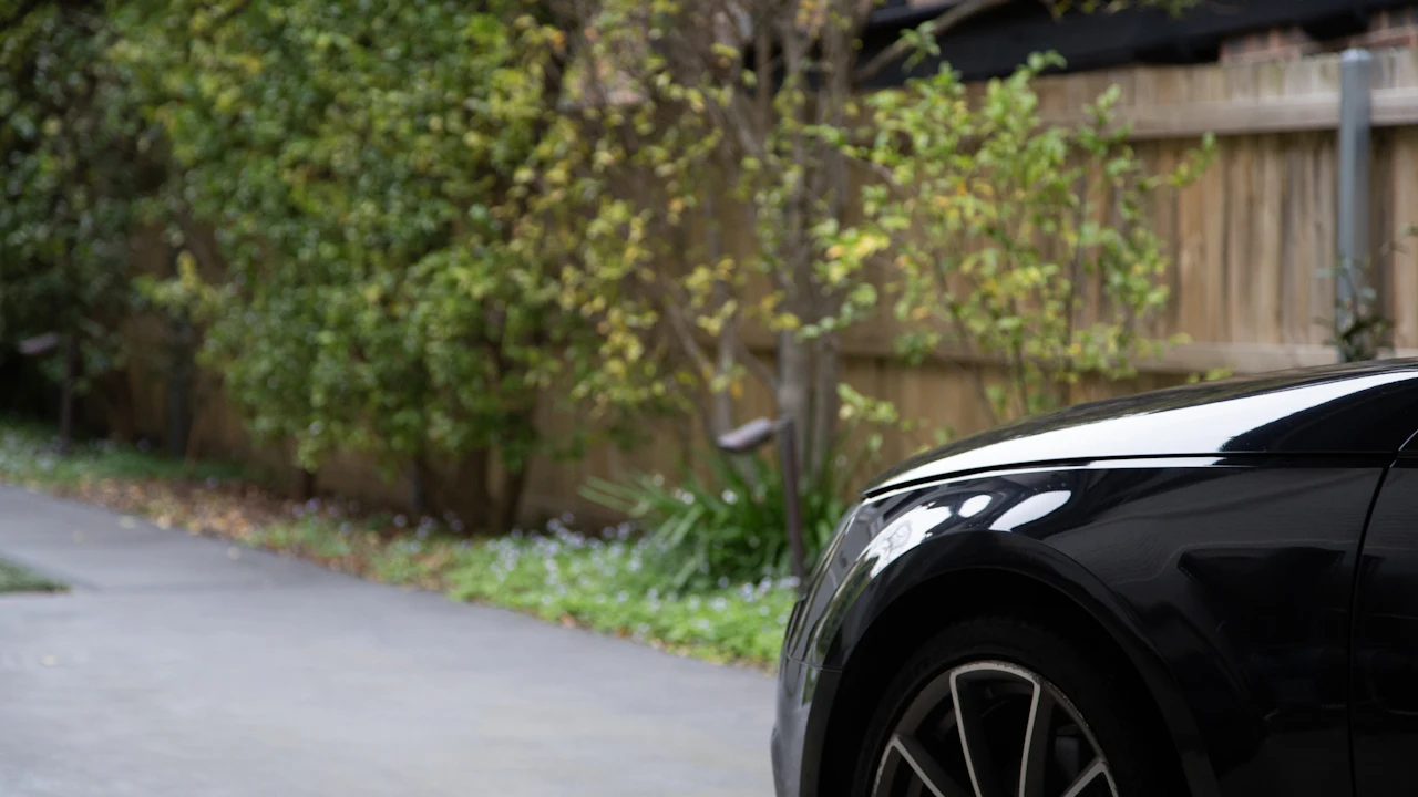 A dark saloon car parked on a residential driveway beside a wooden fence with shrubs behind it