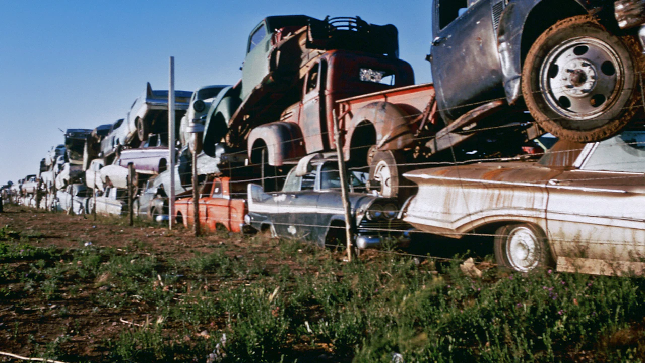 Rows of wrecked cars stacked two high in a scrapyard under a clear sky, with wheels and panels missing