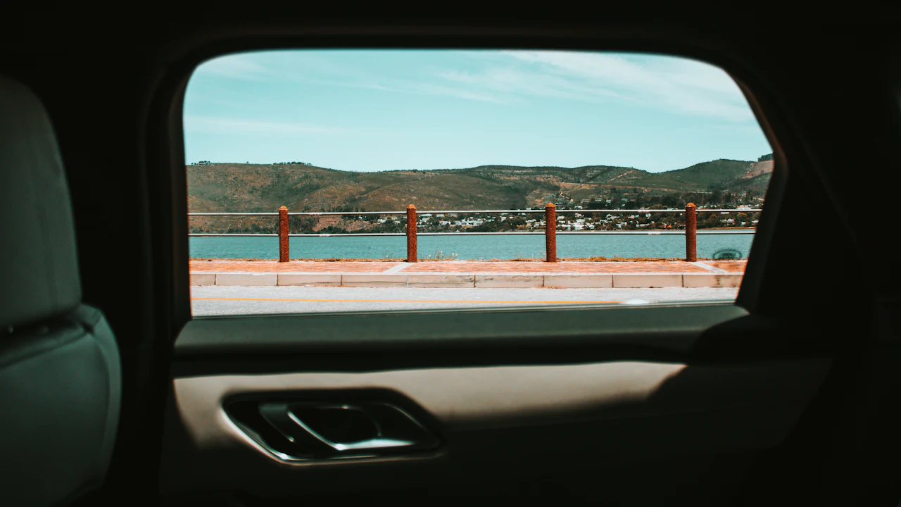The inside of a car's rear door, looking out through its window at a seafront railing, water and hills, with the door's grey trim panel and chrome handle below.