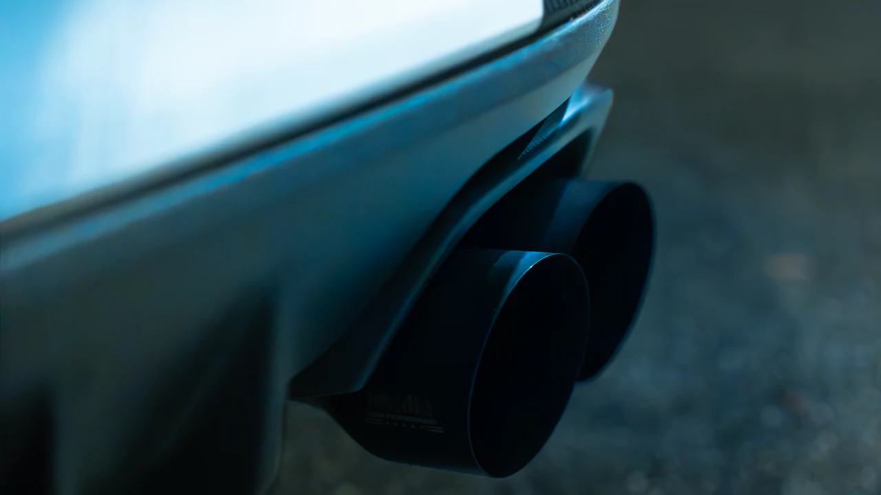 A close-up of the rear bumper of a pale blue car in low light, with a pair of large dark exhaust tips below it and a blurred road surface behind.