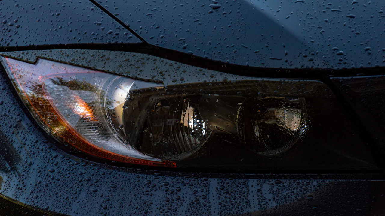 The rain-beaded headlamp, bonnet edge and front bumper of a dark car.