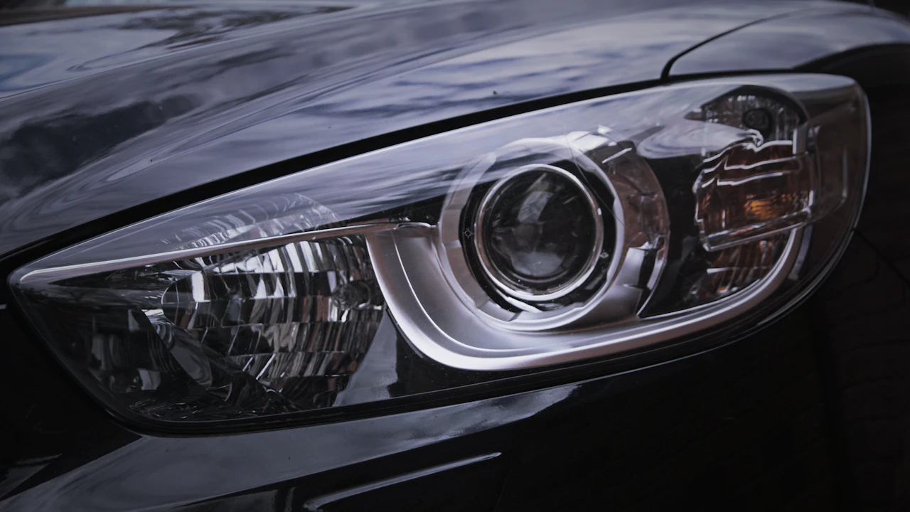 A close-up of the swept-back headlight of a dark, rain-beaded car: a clear lens over a round projector lamp, a chrome surround and an amber indicator, with the glossy bonnet and wing reflecting the sky above it.