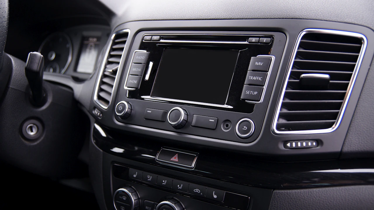A car's centre console with a radio and navigation head unit, its screen, buttons and volume knob, beside a chrome-trimmed air vent.