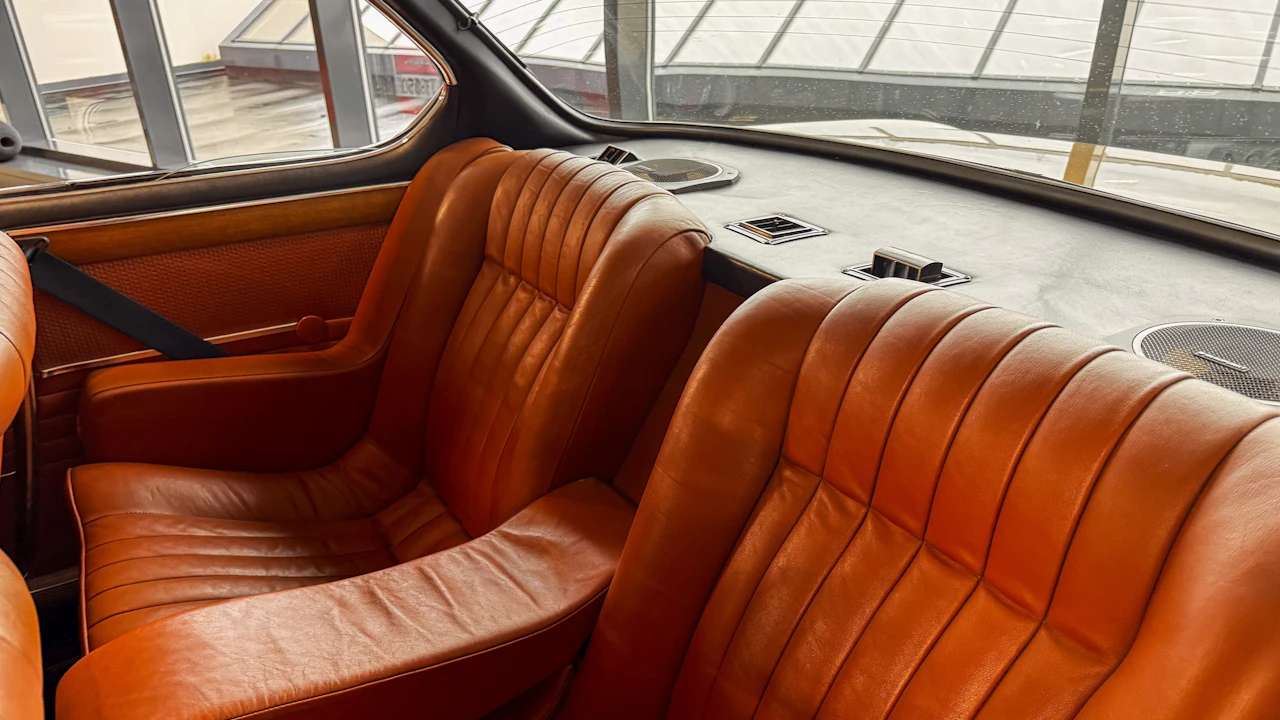 The cabin of a classic car with deeply pleated orange leather front seats, a padded dashboard top with small vents, and a view out through the windscreen to a covered car park.
