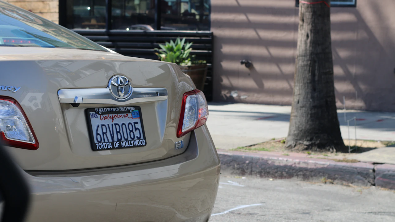 The rear quarter of a gold Toyota saloon parked at a kerb in bright sun, its California plate held in a dealer-branded frame with a coloured renewal sticker in the corner, a palm trunk and a shopfront behind it.
