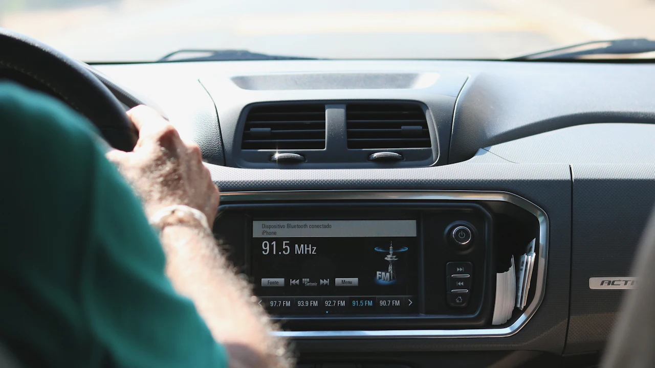 A factory head unit glowing mid-dash, tuned to an FM station, photographed over the driver&rsquo;s shoulder in bright sun