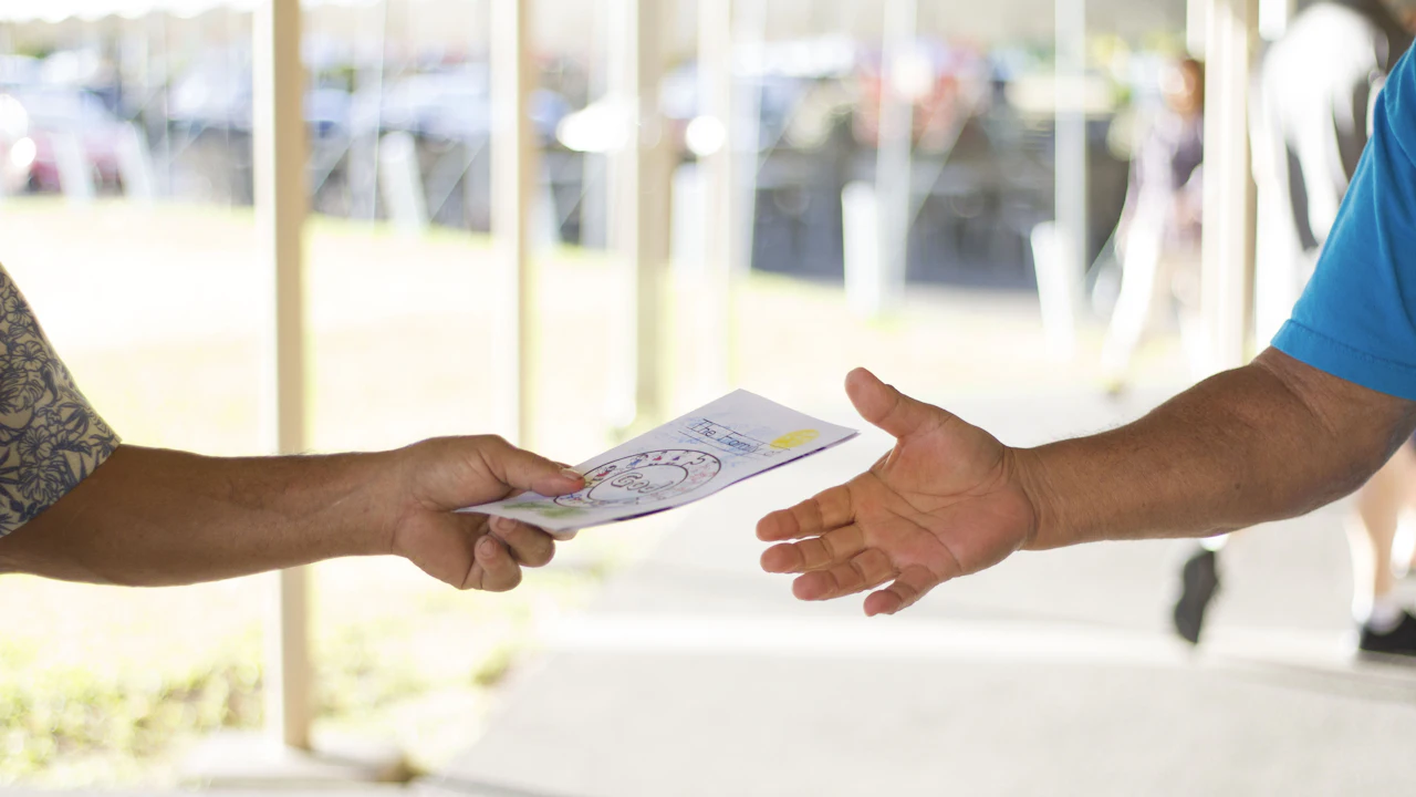 One person handing a folded printed leaflet to another across a low barrier, both arms lit by bright daylight, with parked cars and a marquee out of focus behind them