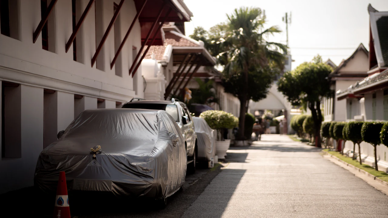 A car parked close against a building under a silver fitted cover, more covered vehicles and a palm tree along the quiet street behind it