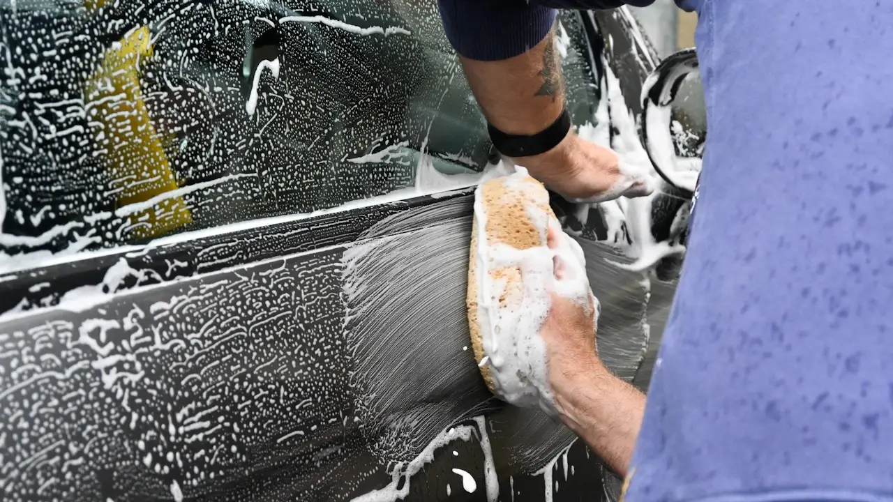Hands washing a dark car door with a soapy sponge.