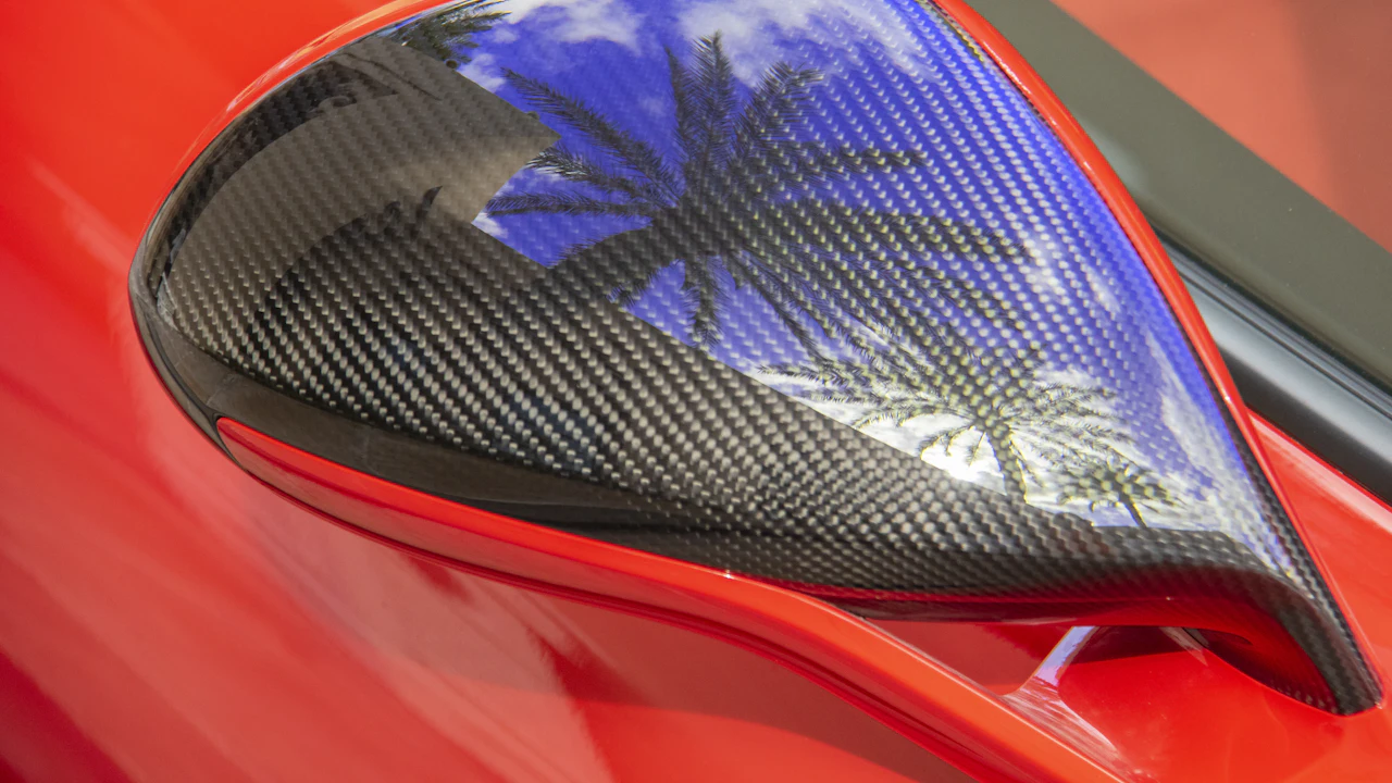 A close-up of a car's carbon-fibre door mirror housing reflecting a palm tree and blue sky, on glossy red bodywork.