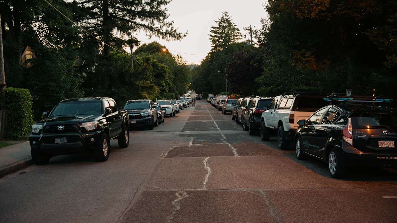 A residential street at dusk lined on both sides with parked cars and pickups beneath overhanging trees