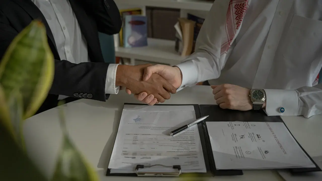 Two people shaking hands over a desk with a clipboard of signed papers.