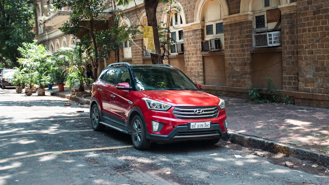 A red compact crossover parked at the kerb of a narrow city street, a colonial-era stone building with arched windows behind it
