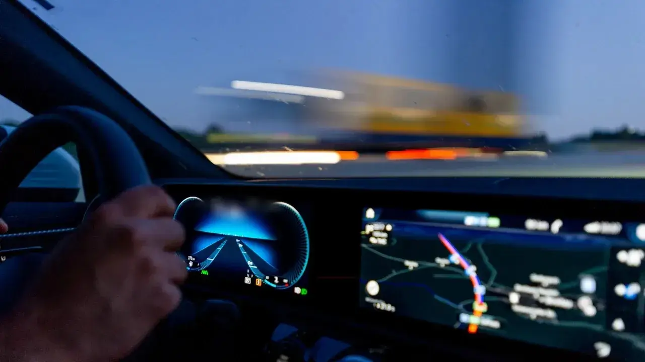 A driver's hand on the steering wheel beside a digital instrument cluster and a wide navigation screen, with a highway blurred through the windscreen.