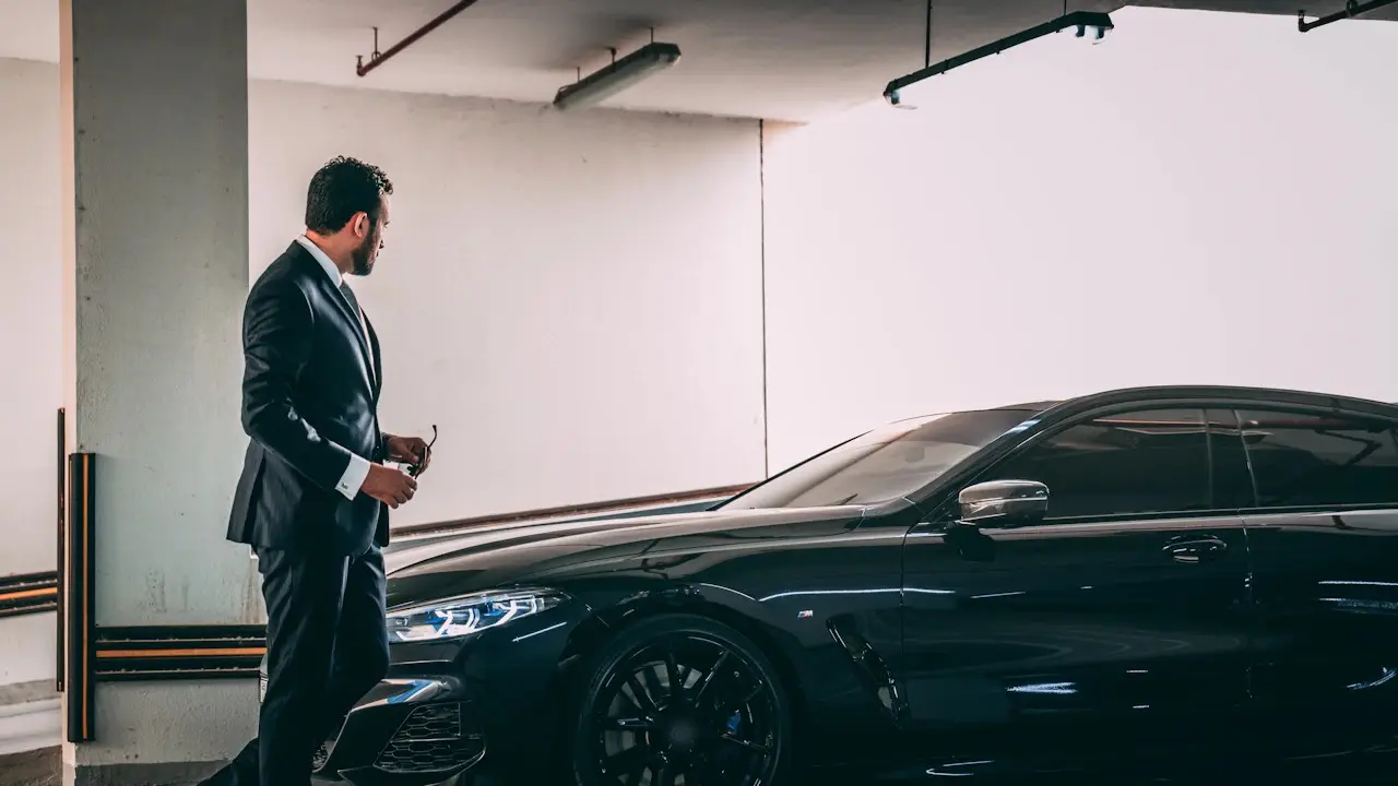 A man in a suit beside a dark luxury coupe in a parking garage.
