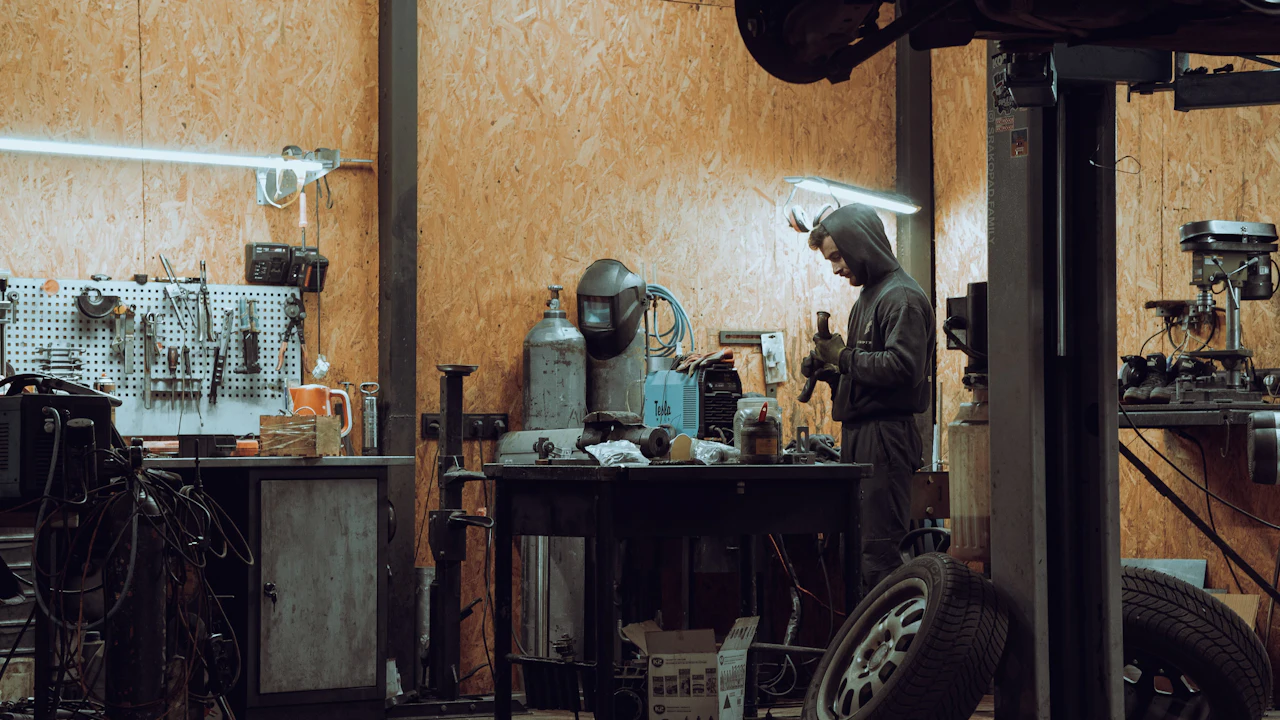 A worker in a home garage workshop beside a raised vehicle and a wall of hand tools