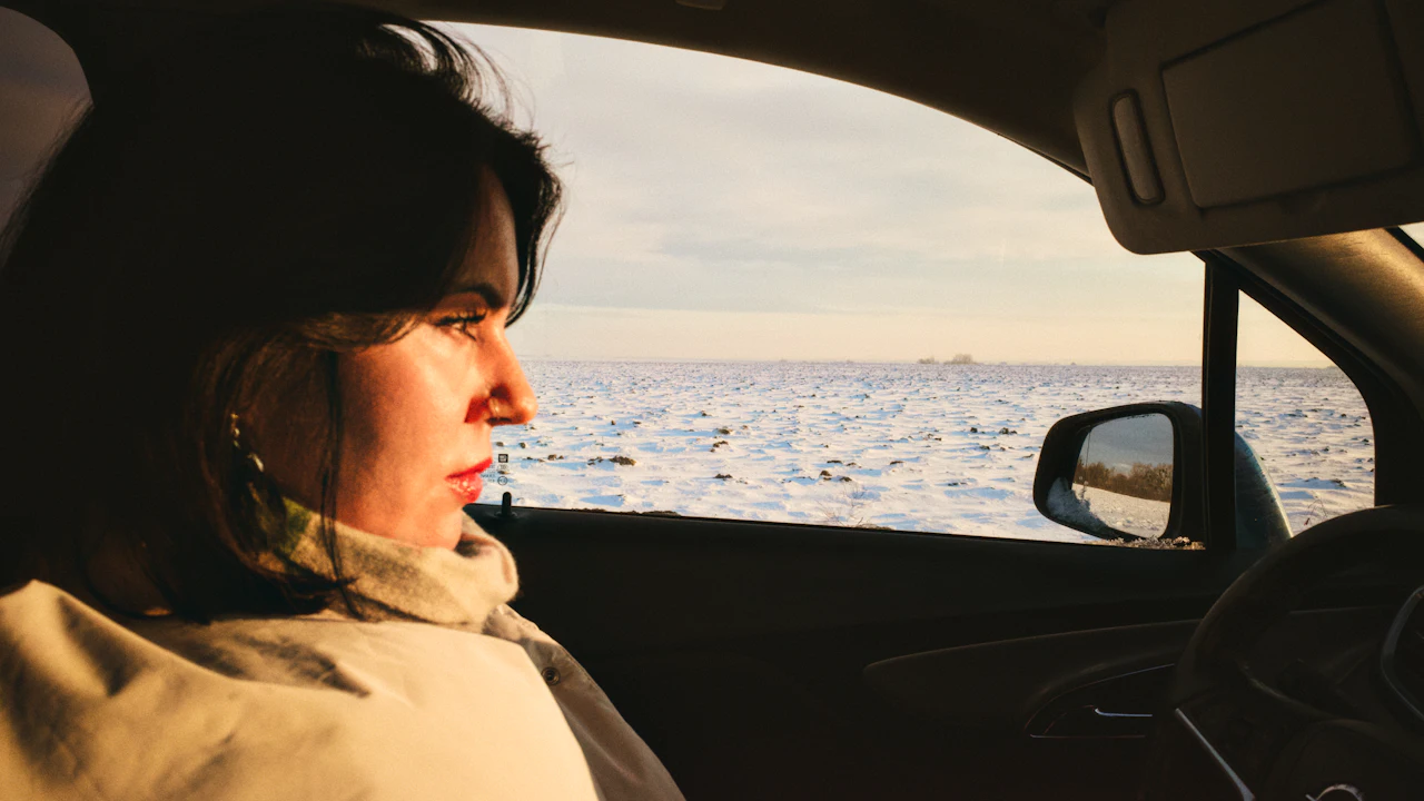 A woman sitting in the passenger seat of a parked car, turned away from the camera and looking out through the open window at a wide expanse of snow and pale winter sky, the wing mirror and door frame dark in the foreground