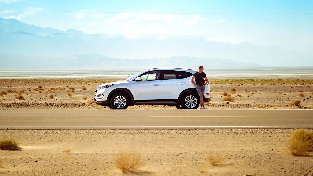A driver leaning on a white SUV parked beside a desert highway.