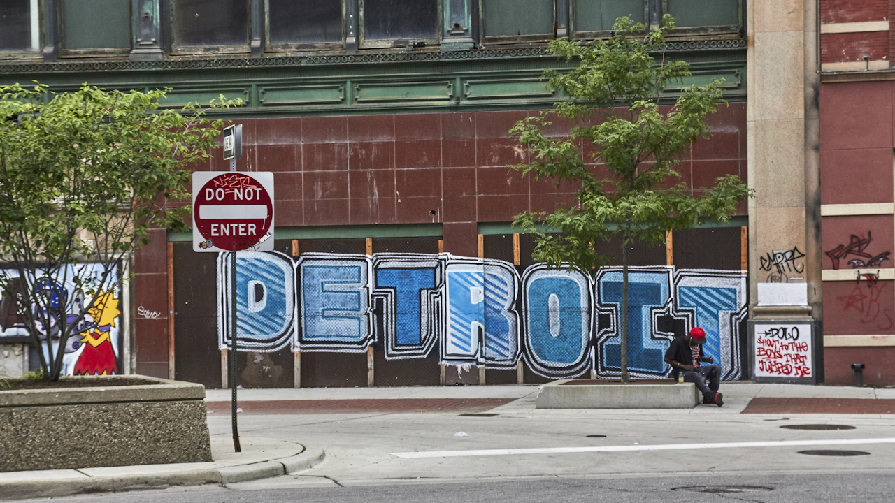 A city street corner with cars parked along it and a large hand-painted DETROIT mural on a boarded facade
